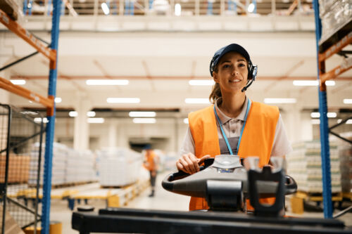 Young female worker driving pallet jack while working at distribution warehouse.